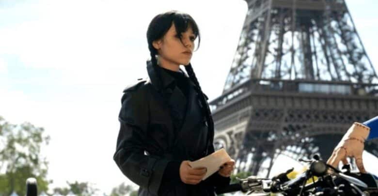 A mysterious woman in a sleek black coat poses with a letter in hand near the iconic Eiffel Tower, while an outstretched arm from the side adds an air of suspense—like a live-action scene you’d find at Universal Studios’ thrilling Netflix experience or a Wednesday-themed attraction, blending cinematic excitement with theme park magic.